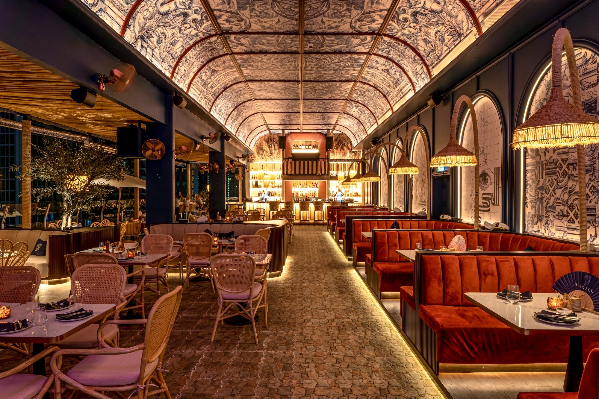 Night time interior of Pastel Bangkok Rooftop Bar and Restaurant, featuring red velvet banquettes, wicker chairs, artistic ceiling decor, and a well-lit bar in the background, creating warm ambiance