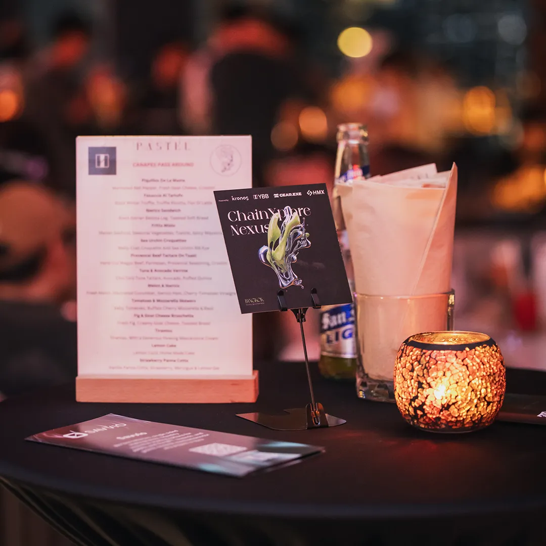 A close-up of a table setup at Pastel Bangkok, featuring a candle, a drink, and event cards for "ChainExplore Nexus," with a softly blurred background of a lively restaurant ambiance.