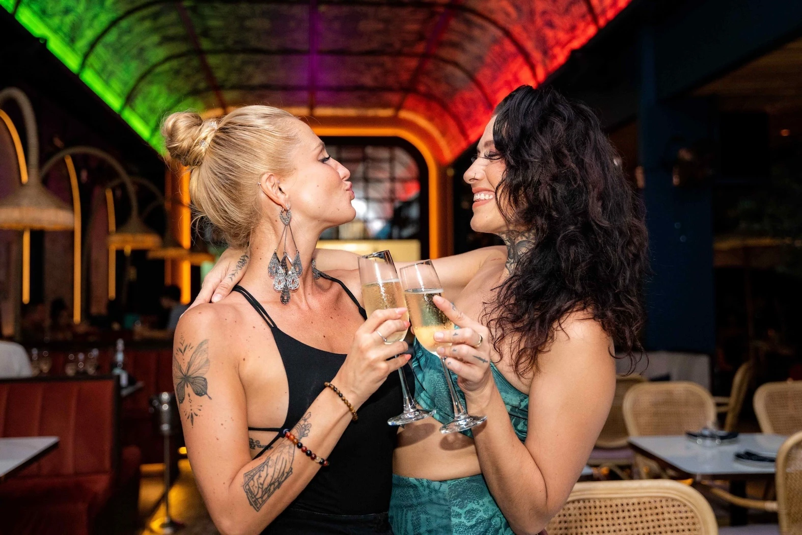 Two beautiful women with tattoos cheerfully clinking champagne glasses at Pastel rooftop bar in Bangkok with colorful ambient lighting in the background.